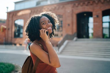 Sırt çantalı Afro-Amerikan kız öğrencinin arka planda kameraya bakıp öğrenme ve öğretim binası girişinde akıllı telefondan sohbet etmesine odaklanın.