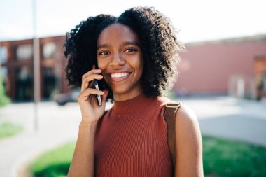 Sırt çantalı mutlu Afrikalı Amerikalı kız öğrenciye odaklanın. Arka planda kampüs binalarıyla akıllı telefondan sohbet ederken kameraya bakın.