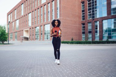 Neşeli, Afro-Amerikan kıvırcık saçlı, kolsuz tişörtlü, akıllı telefonlu, elinde kitaplarla yaz aylarında gezen kadın.