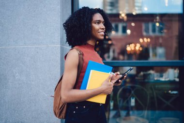 Afro saç stili akıllı telefondan mesaj atan ve elinde not defterleri olan Afro-Amerikan genç kız öğrencinin yan görüntüsü.