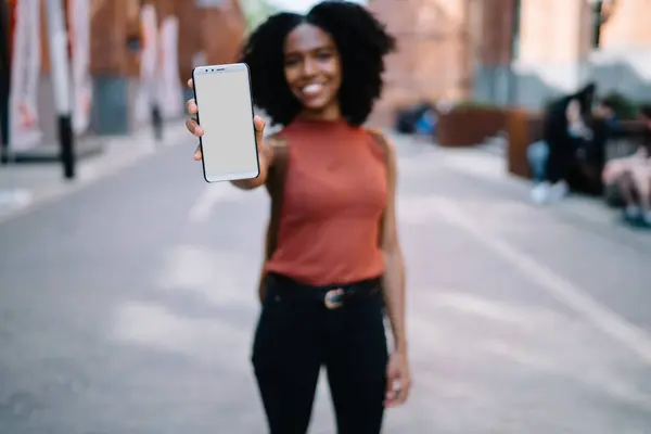 Cheerful black smiling female with Afro hairstyle in casual wear showing blank white screen of cellphone spending time in street of town