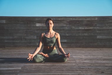 İnce, gevşek, Lotus pozisyonunda dişi yogi elleri dizlerinin üstünde ve gün doğumunda ahşap patikada kapalı gözleri var.