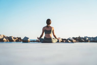Yoga kıyafetleri içinde tanınmayan bir kadın Lotus pozisyonunda meditasyon yaparken sabah su kenarında taş iskelede oturuyor.