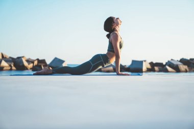 Spor kıyafetli genç sağlıklı bir kadının, arka planda taşlarla deniz kenarında mavi bir minder üzerinde kobra pozisyonunda yoga yaparken yan görüntüsü.