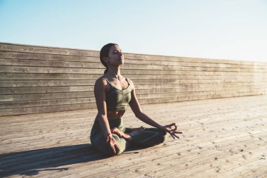 Yoğunlaşmış dingin etnik kadın yogi nilüfer pozisyonunda dizlerinin üzerinde elleri ve gün doğumunda ahşap patikada gözleri kapalı