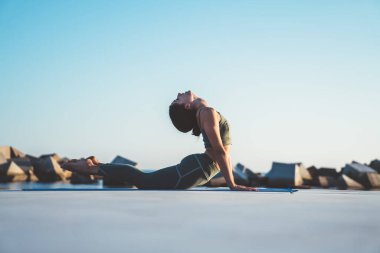 Spor kıyafetli genç güzel bir kadının deniz ve taşların önündeki mavi minderde kobra pozisyonunda yoga yaparken yan görüntüsü. 