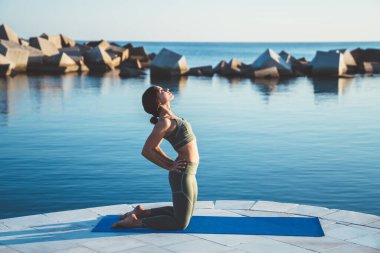 Mavi paspasın üzerinde, elleri kalçasında, barajın kenarında yoga yapan konsantre bir kadının yan görüntüsü.