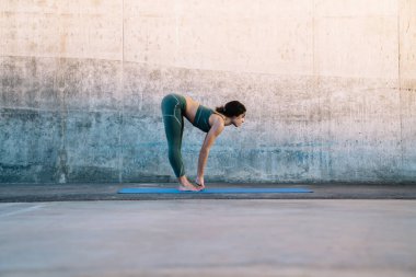 Spor elbiseli genç esmerin yan görünüşü ve yarı yol kaldırma yoga pozu beton duvara karşı fitness paspası üzerinde