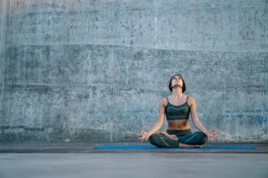 Rahatlatıcı, esnek, spor kıyafetli yetişkin bir kadın yoga minderinde nilüfer pozisyonunda oturuyor ve beton duvara bakıp meditasyon yapıyor.