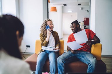 Gündelik giyinen bir kadın akıllı telefondan iş birliği projesinde çalışıyor iş arkadaşlarıyla ofiste koltukta oturuyor.