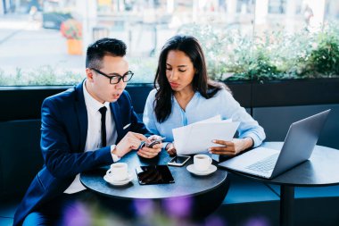 Zarif giyinen, ekrana odaklanan ve elinde kahve ve modern kafede kağıtları incelerken akıllı telefon kullanan yetişkin çoklu etnik girişimcilerin yüksek açısı