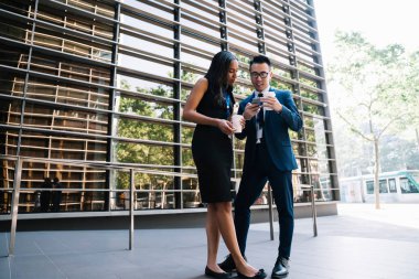 Sıcak içecekle resmi giyinmiş çok ırklı iş arkadaşları olarak ekranlara ilgiyle odaklanmak ve akıllı telefonla etkileşime girmek için çağdaş gökdelen ofis binasının dış yüzeyine karşı ayakta duruyorlar.