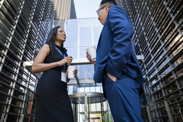 From below of bright woman in formal wear drinking cup of coffee communicating with male with hand in pocket standing in street of business district