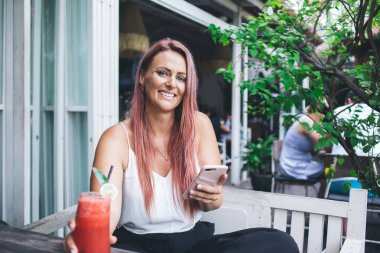 Güzel, uzun saçlı, akıllı telefon ve smoothie ile terasta bankta oturan yaz günü kameraya gülümseyen bir kadın.