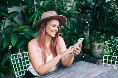 Yazlık kıyafetli, pembe şapkalı, neşeli genç bir kadın tropikal yeşil bitkinin arka planındaki kafede akıllı telefon sörfü yapıyor. 
