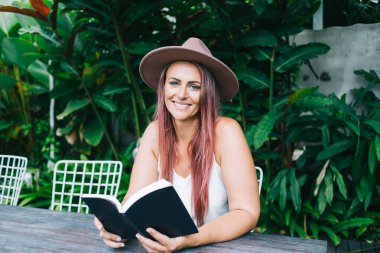 Şapkalı ve boyalı saçlarıyla güzel, neşeli bir kadın masada oturmuş kameraya bakıyor.