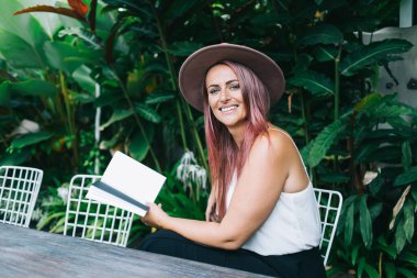 Şapkalı ve boyalı saçlarıyla güzel bir kadın masada oturmuş kameraya bakıyor.