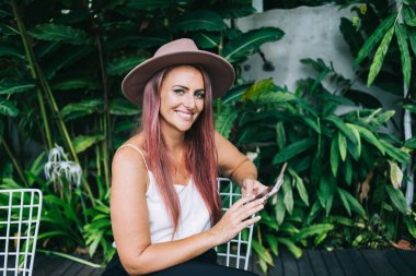 Gülümseyen, bronz tenli, şapkalı, akıllı telefon kullanan ve tatilde tropik bahçede kameraya bakan bir kadın.