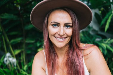 Çekici, mavi gözlü, şapkalı, pembe saçlı, dışarıda yeşil arka planda oturan ve kameraya bakan güzel bir kadın.