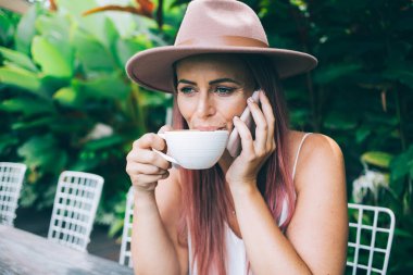 Olumlu gülümseyen, şapkalı, akıllı telefonlu, kahve içen, tropikal yaz gününde açık terasta oturan modern bir kadın.