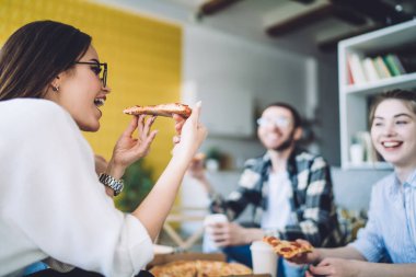 Keyifli genç bir kadın, evdeki sıcak oturma odasında arkadaşlarıyla eğlenirken lezzetli bir pizzayı ısırıyor.