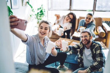 Bir grup mutlu erkek ve kadın ellerinde hazır içecek bardakları ve evde parti sırasında gülümseyerek selfie çekiyorlar.