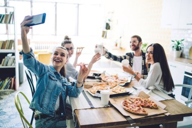 Rahat bir hayat süren neşeli arkadaşlar pizza yer ve bira içerken modern mutfakta dinlenir ve gün ışığında selfie çekerler.