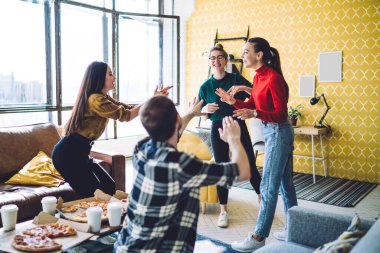 Modern bir dairede pizza kutuları ve tek kullanımlık bardaklarla masaya otururken günlük kıyafetlerle konuşan ve el kol hareketi yapan mutlu insanlar.