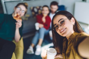 Smiling happy woman in glasses relaxing with people in casual outfit eating pizza and drinking from disposable cup in sunny apartment clipart