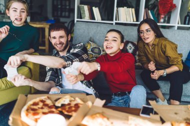 Moda kıyafetleri içinde mutlu arkadaşlar video oyunu oynarken rahat bir kanepede dinleniyor ve modern çatı katında lezzetli pizzalar yiyor.
