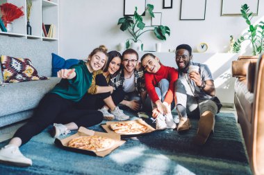 Pizza ve paket servis içkileriyle donanmış mutlu çok ırklı arkadaşlar yerde oturup ev partisi sırasında kameraya bakıyor.