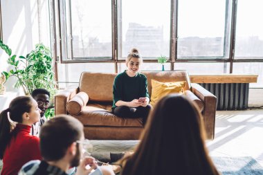 Güzel, genç bir bayan kanepede oturmuş pencereye gülümsüyor ve cep telefonundan mesajları kontrol ederken arkadaşlarıyla konuşuyor.
