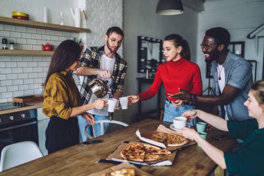 Neşeli, çok ırklı insanlara çay dolduran bir adam açık bir apartmanın mutfağındaki paket pizza kutularıyla masada dikiliyor.