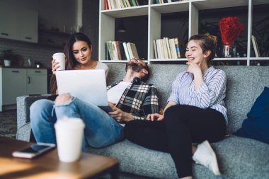 Sakallı sakallı adam şakaya gülüyor ve dizlerinin üstünde dizüstü bilgisayarını tutuyor ve plastik bardaklı neşeli siyah saçlı kadın ile dişlek gülüşlü genç kadın arasında oturuyor.