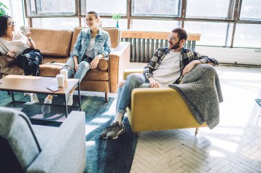 Neşeli genç arkadaşlar kanepede ve koltukta oturuyor oturma odasında dinleniyor, gülümsüyor ve gündüz kahvelerini içiyorlar.