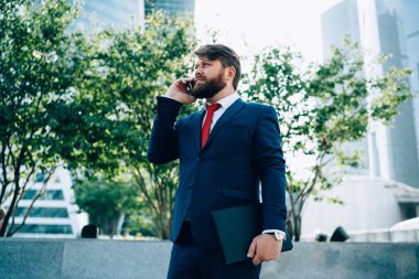 Takım elbiseli ve kırmızı kravatlı dalgın bir işadamı ofis binasının önünde elinde dosyayla dikilip telefonla konuşuyor.