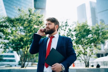 Takım elbiseli, kırmızı kravatlı ciddi sakallı bir iş adamı iş merkezinin yanında elinde dosyayla dikilip telefonla konuşurken başka yöne bakıyor.