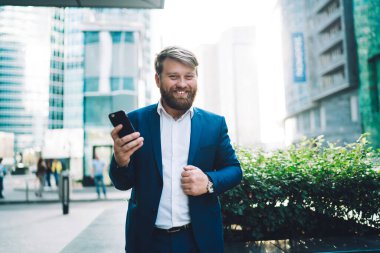 Modayı takip eden iş kıyafetleri içinde memnun bir erkek ofis çalışanı cep telefonuyla mesaj yazarken gülümsüyor ve Business Street 'te kameraya bakıyor. 