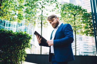 Yeşil ağaçlar ve ofis binaları arka planda dururken tablet kullanan resmi mavi takım elbiseli kendine güvenen modern sakallı işadamlarından. 