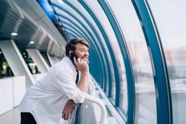 Yandan gülümseyen sakallı adamın günlük kıyafetleri içinde çite yaslanmış cep telefonuyla konuşan hafif geometrik koridorda pencereye bakarken görüntüsü