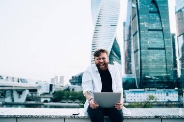 Gözlüklü neşeli hipster adamın portresi. Şehir merkezinde oturmuş, keyifli ve heyecanlı bir erkek programcının yeni bilgisayarında uzaktan çalışmanın keyfini çıkarırken mutlu bir şekilde dizüstü bilgisayar geliştiriyor.