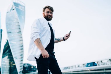 Modern şehirde aynalı gökdelenli sörf yapan, saat takan, beyaz tişörtlü, neşeli, sakallı bir erkek.  