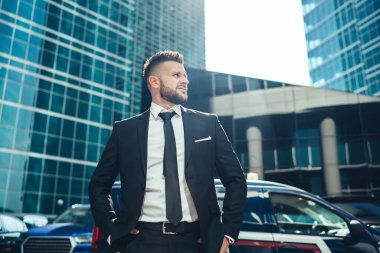 Siyah takım elbiseli zarif sakallı genç adam. Elleri belinde, modern şehrin güneşli sokaklarında partneriyle buluşmayı bekliyor.