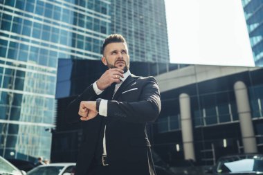 Düşünceli, başarılı iş adamının düşük açısı. Dev modern iş merkezinin önünde duruyor. Bekliyor ve düşünüyor.