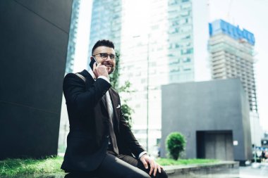 Gözlüklü, şık siyah takım elbiseli, neşeli, kendine güvenen işadamının caddede taş bir bankta otururken yaptığı telefon görüşmesi çağdaş gökdelen binasına karşıydı.