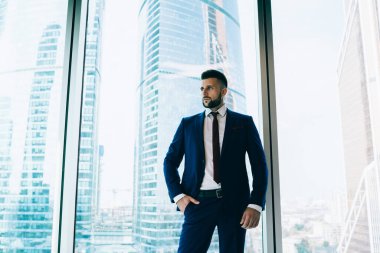 Ofis penceresinin önünde el ele tutuşmuş şık takım elbiseli, konsantre ve ciddi bir erkek işçiden başka tarafa bakan bir ofis penceresi var.