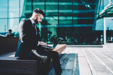 Siyah takım elbiseli genç sakallı işadamının ofis binasının yanındaki bankta oturması ve dizüstü bilgisayardan finansal haberler okuması.