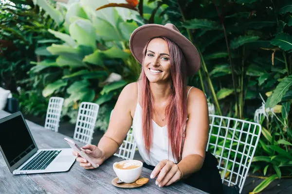 Bright freelancer with dyed hair sitting in garden with cup of aromatic coffee on table and browsing social media during break while looking at camera