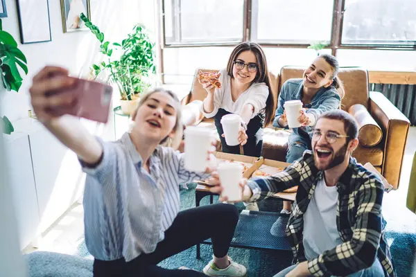 Group of excited young people with cups of drinks to go taking selfie while sitting on sofas and having fun during party