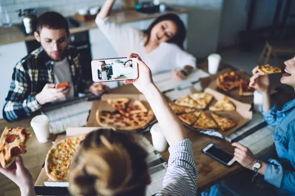 High angle selective focus of unrecognizable female taking picture of cheerful mates having fun at table while eating pizza during friends meeting at home
